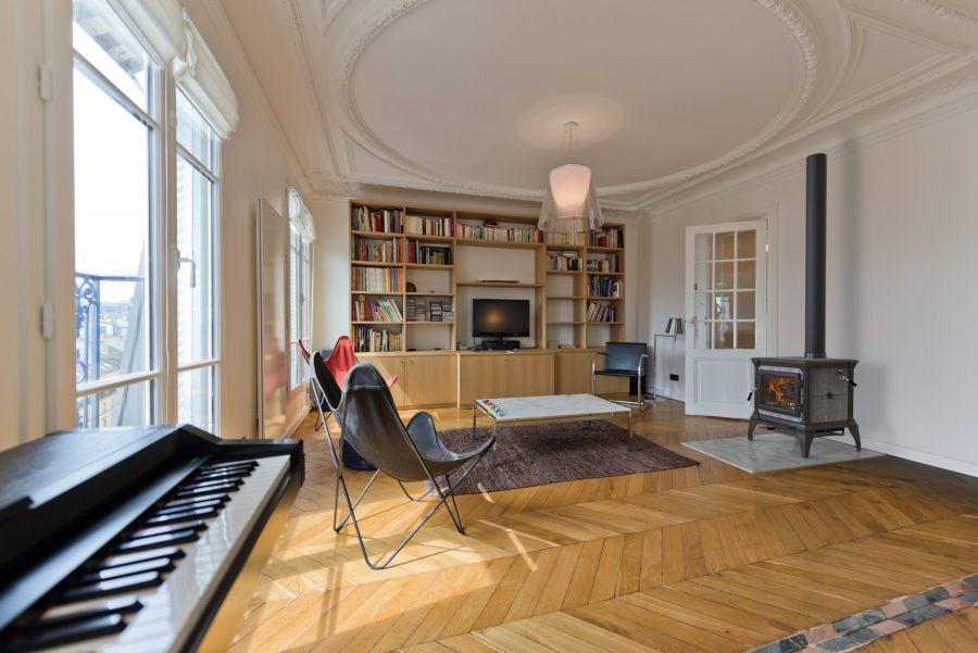 Small black wood fireplace in a open living room with wood floors a piano some large windows and some seating