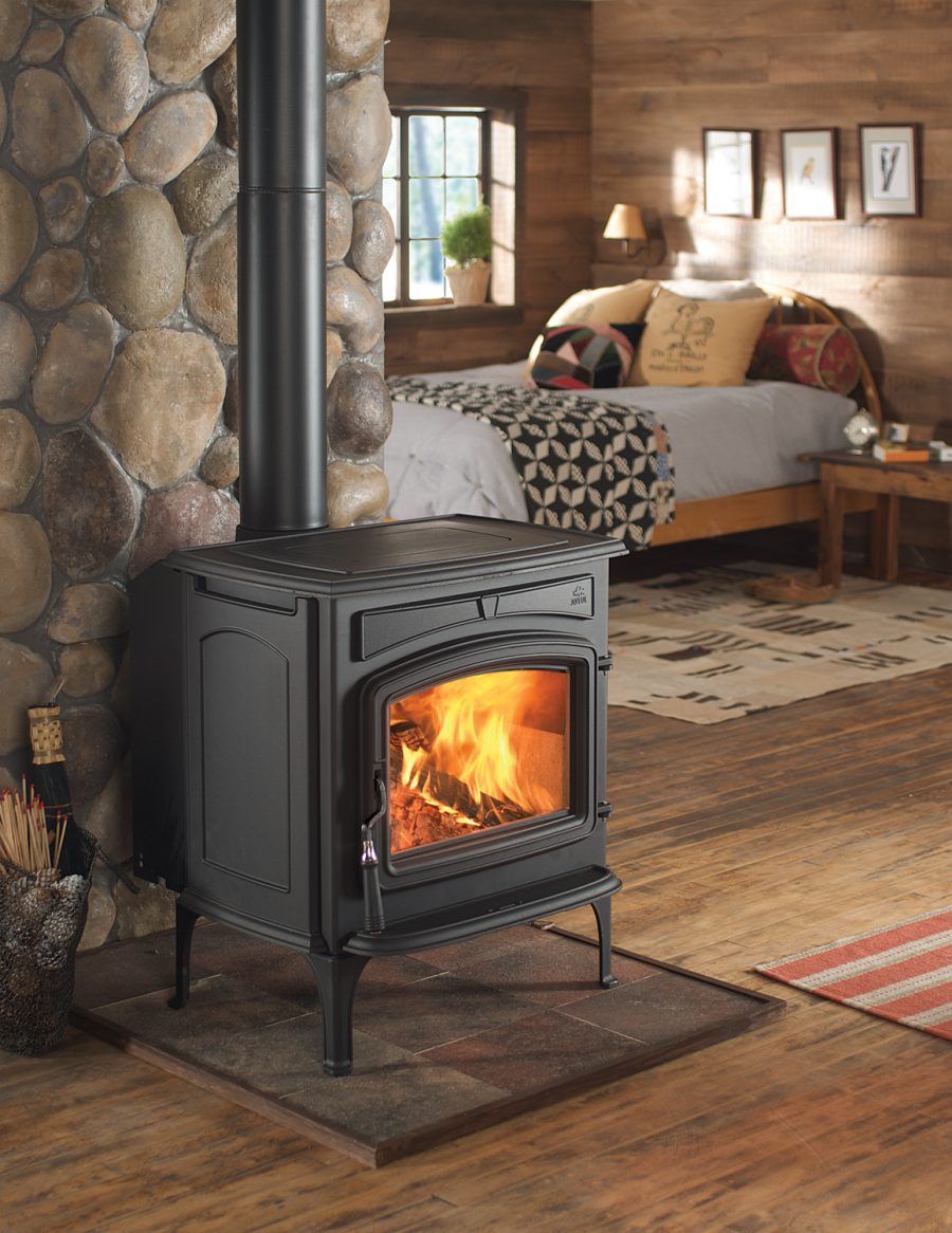 Black wood fireplace in the bedroom of a cabin against a stone chimney with a bed in the background