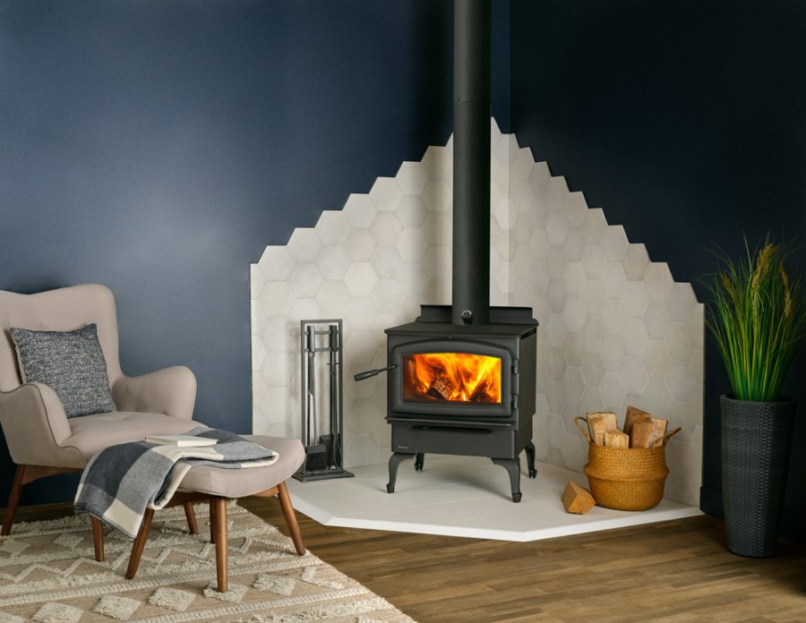 Black woodstove in the corner of a living room with white tile surrounding it and a small seating area next to it