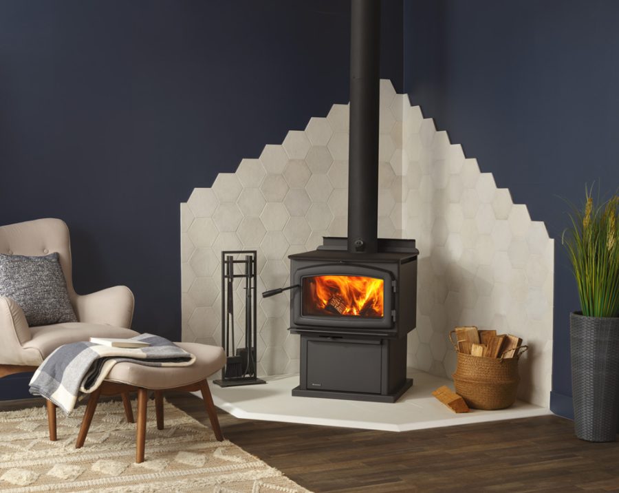 Black woodstove in the corner of a living room with white tiles lining the walls around it with a warm fire going and a small seating area next to it