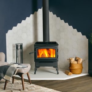 Black woodstove with a fire going in the corner of a room with white tiles lining the blue walls around it with a seating area