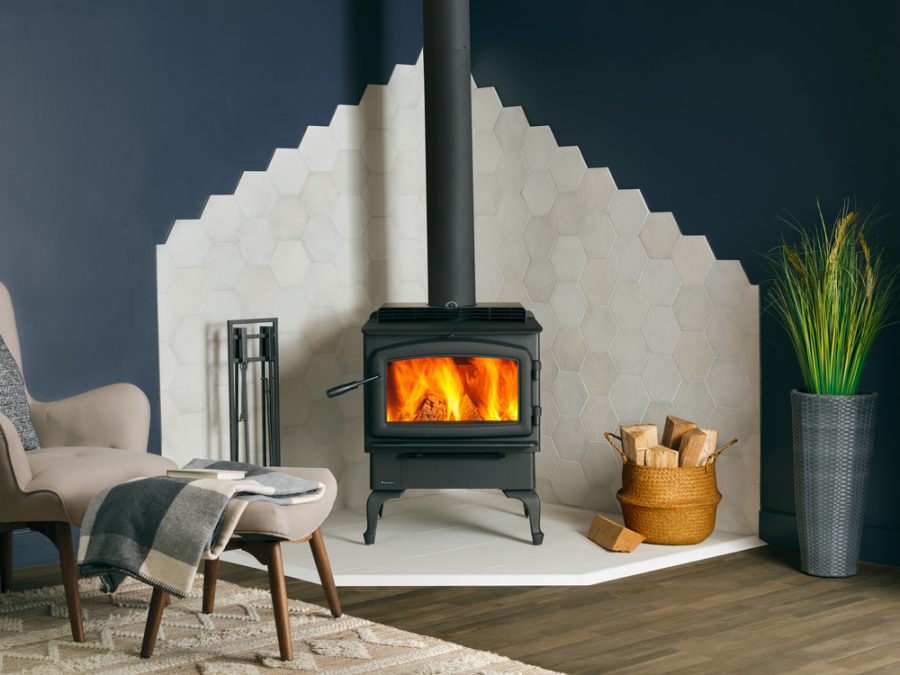 Black woodstove with a fire going in the corner of a room with white tiles lining the blue walls around it with a seating area