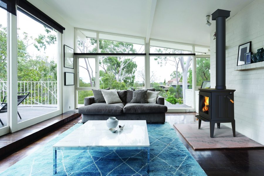 Black wood stove in a living room with blue carpet and large windows covering the walls