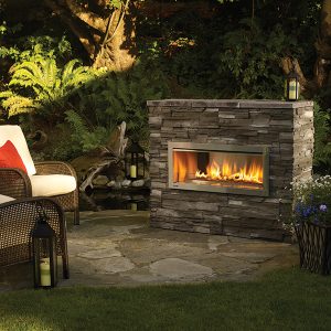 Outdoor fireplace built in a stone seating area with woods in the background and comfortable seating