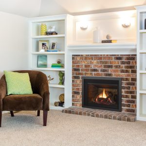 A cozy living room corner featuring a brown chair with a green pillow, built-in white shelves with various decorative items, and a brick fireplace with a burning fire.