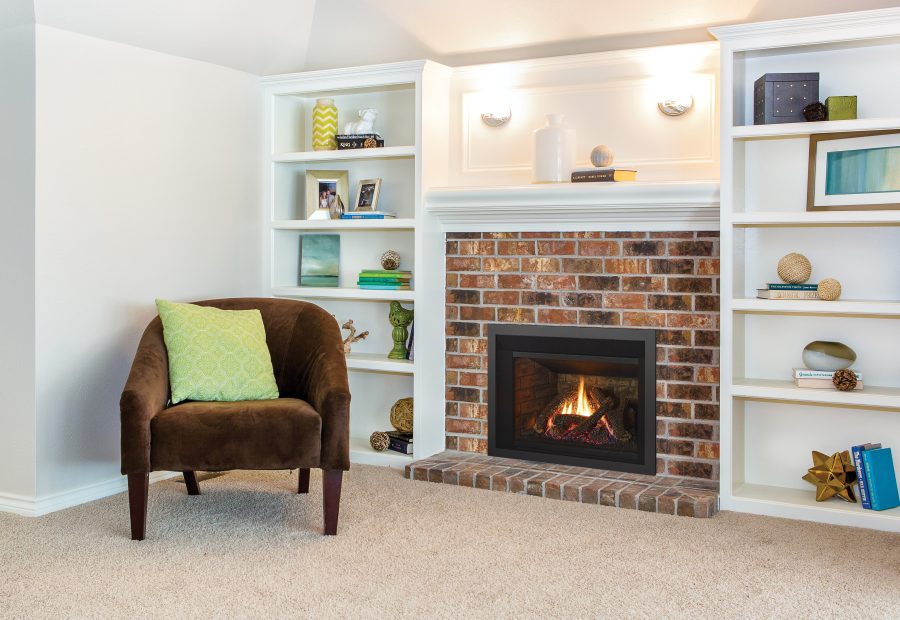 A cozy living room corner featuring a brown chair with a green pillow, built-in white shelves with various decorative items, and a brick fireplace with a burning fire.