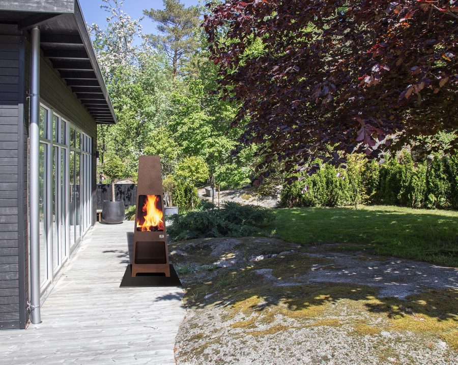 Outdoor pyramid shaped wood fireplace next to a modern home with some woods in the background